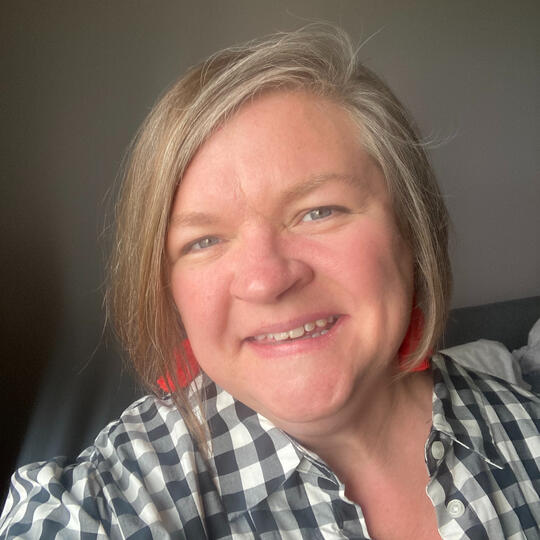 Katie is a white woman in her 40s, chin length light brown and gray hair, and is wearing a black and white shirt over a black top with red earrings